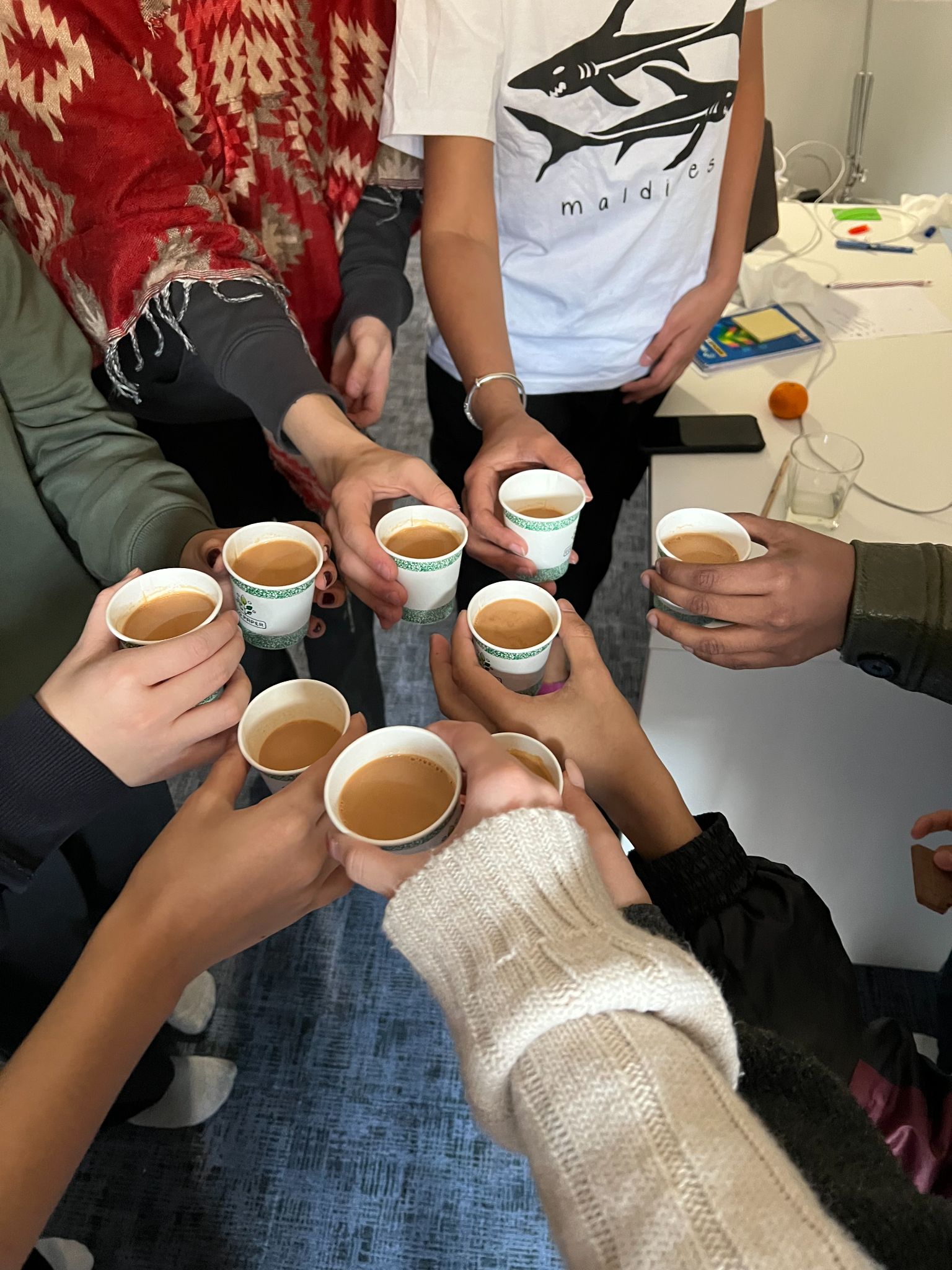A ring of hands raising paper cups of chai over a table